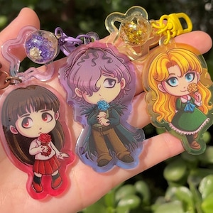 May include: A selection of acrylic keyrings with anime-style character designs. The keyrings showcase various characters, including a girl in a red skirt, a character with purple hair, and a blonde girl in a green dress. Some keyrings have glitter or floral embellishments.