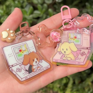 May include: Keychains featuring cartoon dogs inside a handheld gaming device. One keychain has a brown dog, the other a yellow dog. Additional charms include a paw print and a star. The keychains have pink and brown accents.
