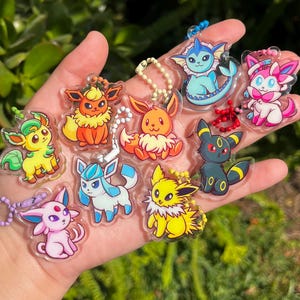 Puede incluir: Una colección de llaveros acrílicos coloridos con varios personajes de dibujos animados. Los llaveros muestran una variedad de diseños, incluyendo criaturas naranjas, amarillas, azules, rosas y verdes. Cada llavero tiene una cadena plateada.
