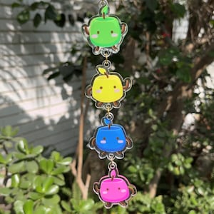 Puede incluir: Un colgante decorativo con cuatro colgantes en forma de manzana de colores brillantes. Los colgantes son verdes, amarillos, azules y rosas, cada uno con brazos marrones y una cara sonriente. Están unidos por pequeñas cadenas doradas.