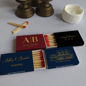 May include: A collection of personalized matchboxes in various colors, including burgundy, navy, and black. Each box features custom text and dates in gold lettering. Some boxes display initials, names, and a house illustration. Matches with red tips are visible.