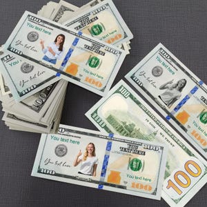 May include: Several custom $100 bills with the text "You text here" and images of people. The bills are stacked and arranged on a gray surface. The bills are white, green, and orange.