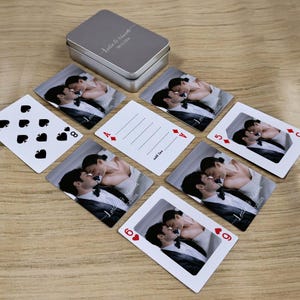 May include: A set of personalized playing cards featuring a wedding photo, with a silver tin case. The cards display a couple kissing, with the names "Leslie & Marcel" and the date "06.12.2026" printed on them. Some cards show traditional suit symbols.
