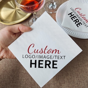 May include: White square napkin with the text "Custom LOGO/IMAGE/TEXT HERE" in burgundy and black. The napkin is held by a hand, with a cocktail glass and a plate with a napkin in the background. The napkin is made of a textured paper material.