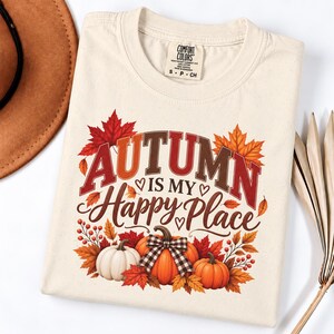 Peut inclure: Un t-shirt couleur crème avec un motif portant l'inscription "AUTUMN IS MY Happy Place" en lettres rouges et brunes. Le design comprend des citrouilles orange et blanches, dont une avec un nœud à carreaux noir et blanc, entourées de feuilles.