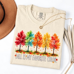 Peut inclure: Un t-shirt à manches courtes couleur crème avec un motif aquarelle représentant cinq arbres aux teintes rouge, verte, orange, jaune et rouge orangé. Sous les arbres, le texte FALL IS MY FAVORITE COLOR est écrit en noir avec un contour.