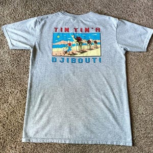 May include: A heather gray t-shirt featuring a colorful graphic of camels and people in a desert landscape. The text "TIN TIN A DJIBOUTI" is printed above and below the graphic in red and blue.