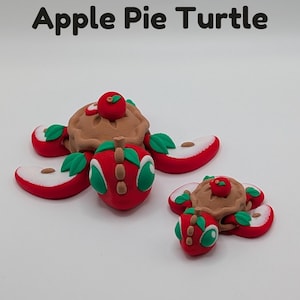 May include: Two whimsical clay turtle figurines, each with a pie crust body and apple slice limbs. The turtles have red apple heads with green leaves and a small apple on top. The text "Apple Pie Turtle" is at the top.