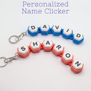 May include: Two personalized name clickers. One is blue with the name "DAVID" and the other is coral with the name "SHARON". Each name is spelled out with individual letter tiles. The text "Personalized Name Clicker" is at the top.
