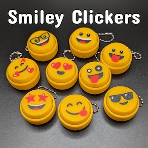 May include: A collection of yellow Smiley Clickers with various emoji faces, including hearts, sunglasses, and a tongue sticking out. Each clicker has a silver keychain attached. The text "Smiley Clickers" is at the top.