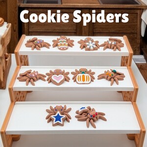 May include: Brown cookie spiders with colorful decorations, displayed on a tiered wooden shelf. The cookies have various designs, including hearts, stars, and geometric patterns. The text "Cookie Spiders" is at the top.