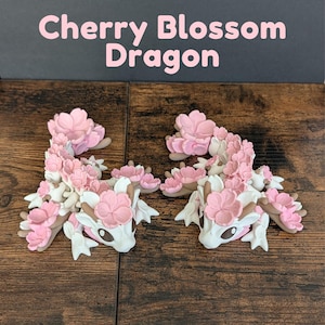 May include: Two dragon figurines with cherry blossom accents. The dragons are white with pink and brown details, and the text "Cherry Blossom Dragon" is above them. The figurines are on a wooden surface.