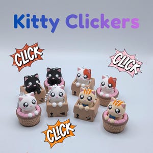 May include: A collection of colorful cat-shaped clickers, each nestled in a miniature basket or cardboard box. The clickers feature various colors and patterns, with the text "Kitty Clickers" displayed above.