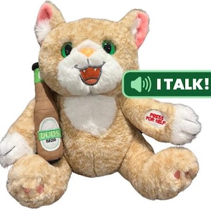 May include: A plush, tan-colored cat toy with green eyes and a smiling mouth, holding a beer bottle labeled "Duds Beer." The toy has a green button that says "I TALK!" and a paw with the text "PRESS FOR HELP."