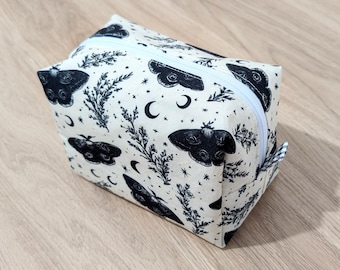 May include: A cream-colored zippered pouch featuring a repeating pattern of black moths, crescent moons, and botanical elements. The rectangular bag is made of fabric and sits on a wooden surface. The zipper is white.