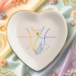May include: Heart-shaped white ceramic trinket dish with a gold rim. The dish features a rainbow-colored illustration of two hands holding, with the names "Sophia" and "Emma" and the date "04/08/26". The dish is surrounded by jewelry and flowers.