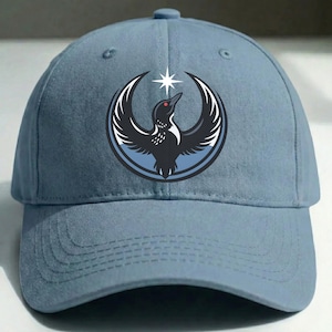 May include: A light blue baseball cap with a circular emblem. The emblem features a black bird design, a white star, and a blue background. The bird has a red eye and is surrounded by white and black curved lines.