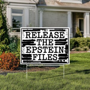 May include: A white yard sign with black text that reads "RELEASE THE EPSTEIN FILES." The sign is in front of a house with a green lawn and landscaping.