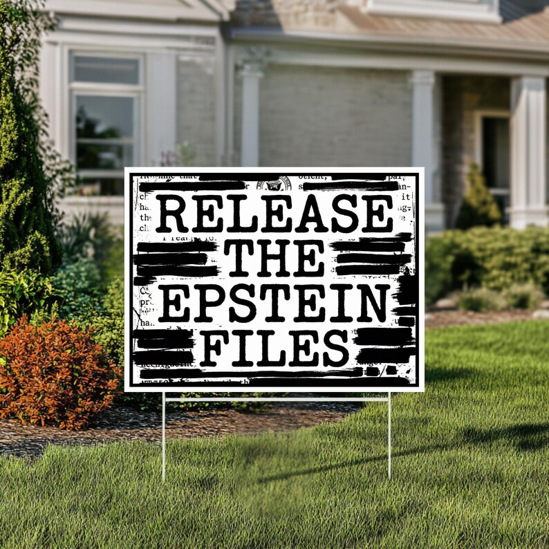 May include: A white yard sign with black text that reads "RELEASE THE EPSTEIN FILES." The sign is in front of a house with a green lawn and landscaping.