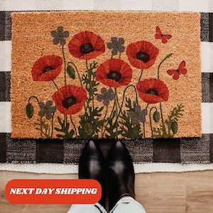 May include: Rectangular doormat with a floral design featuring red poppies and purple flowers. Two red butterflies are also depicted. The doormat is made of a natural fiber material and is placed on a black and white checkered rug.