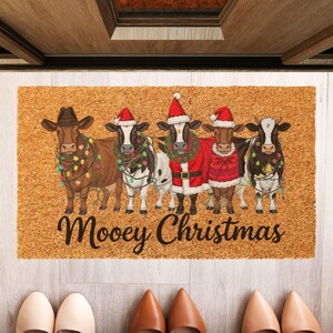 May include: A rectangular doormat with a light brown background features five cartoon cows dressed for Christmas. The cows wear Santa hats, festive outfits, and string lights. The words "Moovey Christmas" are written in a script font.