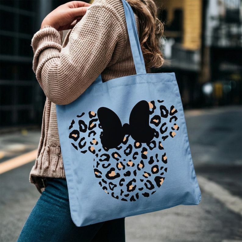 Puede incluir: Bolso tote azul claro con un dise&ntilde;o de Minnie Mouse. El dise&ntilde;o incluye un lazo negro y un estampado de leopardo en tonos azules, negros y beige. El bolso lo lleva una persona.