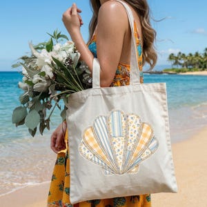 May include: A cream-coloured tote bag with a seashell design. The shell is made of fabric patches in blue, yellow, and white patterns. The bag has a simple design with a beach theme, ideal for carrying shopping or beach items.