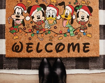 May include: A rectangular doormat featuring Disney characters in Santa hats and scarves, with the word "WELCOME" in black. The characters include Mickey Mouse, Minnie Mouse, Donald Duck, Goofy, and Pluto, all decorated for Christmas. The mat is on a black and white checkered rug.