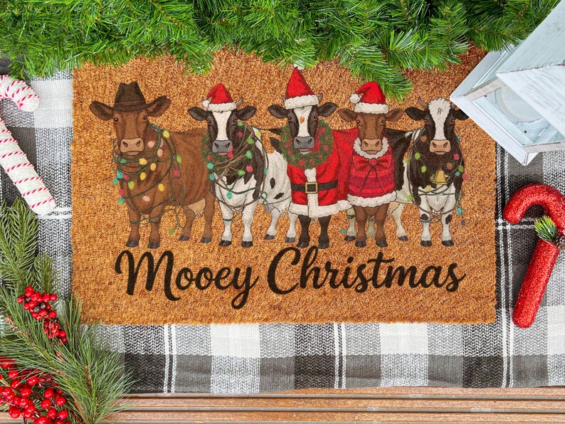May include: A rectangular doormat with a festive design. Five cows are dressed in Christmas attire, including Santa hats and lights. The text "Moovey Christmas" is written across the bottom in a script font. The mat is brown with a textured surface.