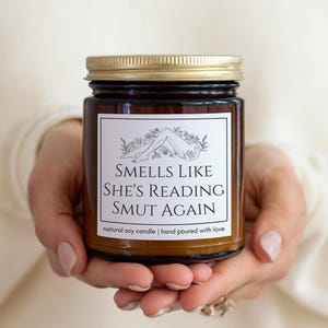 May include: A brown glass jar candle with a gold lid. The label reads "Smells Like She's Reading Smut Again" with a line drawing of legs and flowers. The text below says "natural soy candle | hand poured with love."