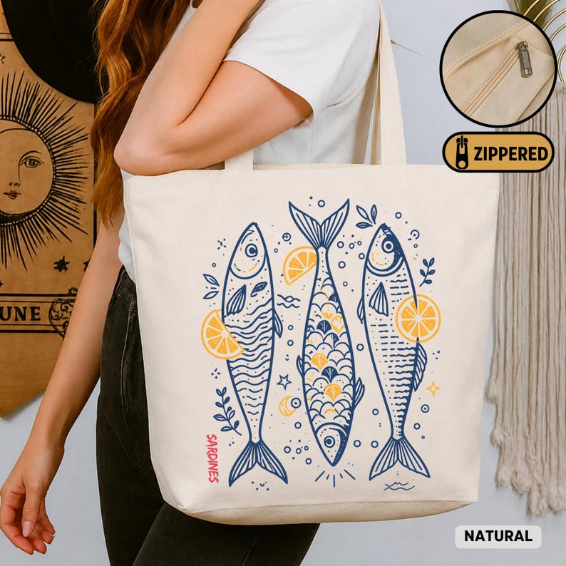 May include: A natural canvas tote bag featuring a blue and yellow illustration of three sardines with lemon slices. The bag has a zippered closure and the word "Sardines" is printed on the side.