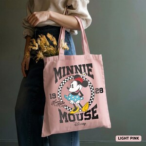 May include: Light pink tote bag featuring Minnie Mouse. The bag has a graphic of Minnie Mouse in a polka dot dress, with the words "MINNIE MOUSE" and "Disney" printed on it. The bag is held by a person.