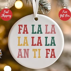 Può includere: Un ornamento natalizio in ceramica bianca con le parole "FA LA LA LA LA LA LA AN TI FA" in lettere rosse, verdi, gialle e rosa. L'ornamento ha un foro nella parte superiore per essere appeso con un nastro bianco. Decorazioni festive natalizie.