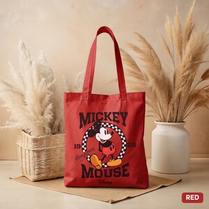 May include: Red tote bag featuring Mickey Mouse. The bag has a black and white graphic of Mickey Mouse in a circle with the words "Mickey Mouse" and "Disney" printed on it. The bag has two red handles.