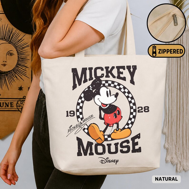 May include: A natural canvas tote bag featuring a vintage Mickey Mouse graphic. The design includes the words "Mickey Mouse" and "Disney" along with the years "19" and "28". The bag has a zippered closure and is made of natural-colored canvas.