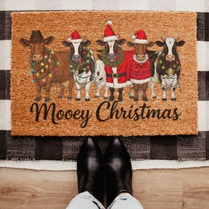 May include: A rectangular doormat with a brown background features five cartoon cows dressed for Christmas. The cows wear Santa hats, lights, and a Santa suit. The text "Moovey Christmas" is written in a script font.
