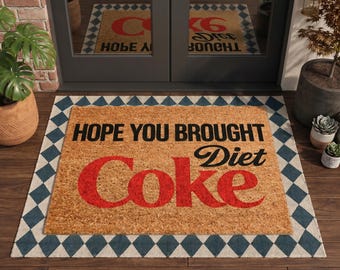 May include: A rectangular tan coir doormat featuring the black text HOPE YOU BROUGHT and the red text Diet Coke. The mat is layered over a larger rectangular mat with a blue and white diamond pattern, positioned in front of double glass doors.