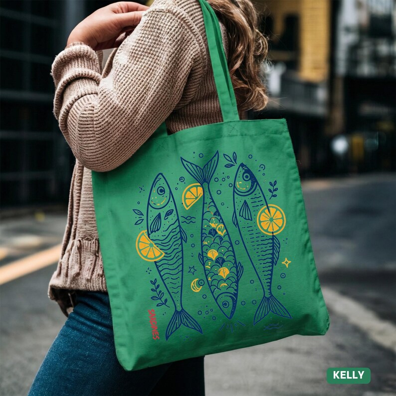 May include: Green tote bag featuring a whimsical design of three fish with lemon slices and botanical elements. The fish are in blue and yellow, with the word "SPARKLING" in red. The bag is carried over the shoulder.