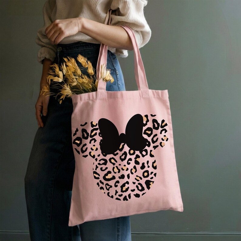 Puede incluir: Un bolso tote rosa con un lazo negro y un dise&ntilde;o de cabeza de rat&oacute;n con estampado de leopardo. El bolso tiene dos asas y est&aacute; hecho de una tela suave. El bolso lo sostiene una persona.
