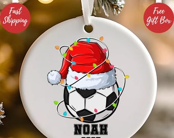 Op de afbeelding: Een witte keramische kerstbal met een voetbalontwerp, een kerstmuts en lichtsnoeren. De bal heeft de naam "NOAH" en het jaar "2025" in zwart gedrukt. Een wit lint is bevestigd om op te hangen.