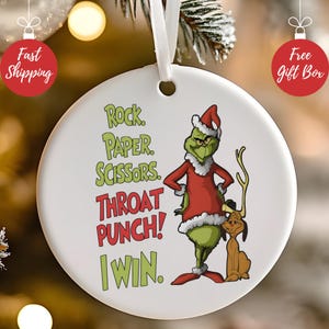 May include: A round white Christmas ornament featuring the Grinch in a Santa outfit, with the text "Rock, Paper, Scissors, Throat Punch! I Win." The Grinch is accompanied by a brown dog-like character.