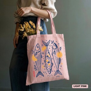 May include: Light pink tote bag featuring three stylized fish illustrations in navy blue with yellow lemon accents. The bag has long handles and the word "Sardines" is printed on the side. The words "Light Pink" are printed on the bottom right.