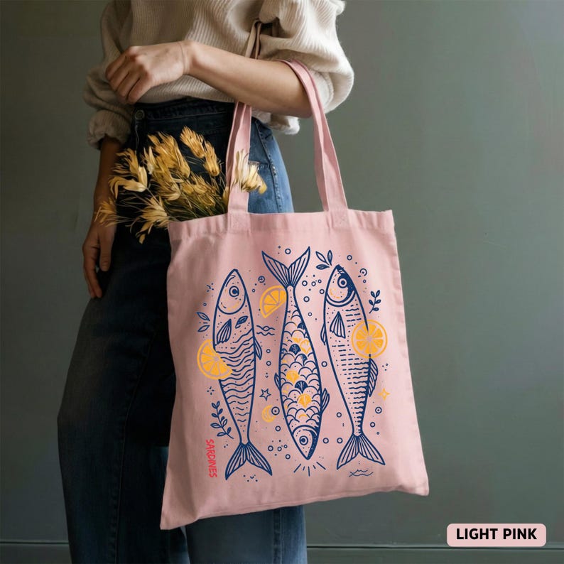 May include: Light pink tote bag featuring three stylized fish illustrations in navy blue with yellow lemon accents. The bag has long handles and the word "Sardines" is printed on the side. The words "Light Pink" are printed on the bottom right.