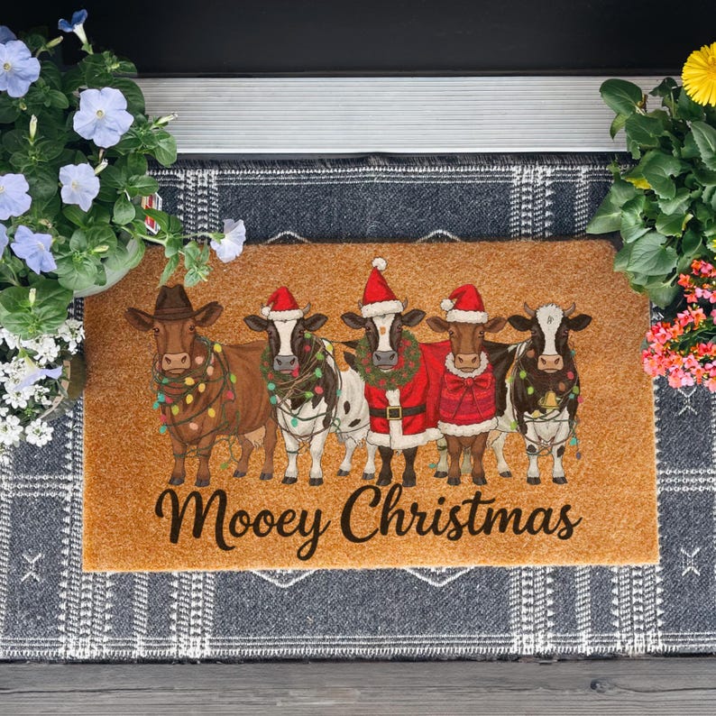 May include: A rectangular doormat with five cartoon cows dressed for Christmas. The cows are brown and white, wearing Santa hats, Christmas lights, and festive outfits. The text "Moovey Christmas" is written in a script font.
