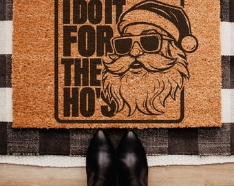 May include: A rectangular doormat featuring the text "I DO IT FOR THE HO'S" and a graphic of Santa Claus wearing sunglasses. The mat is brown with a black border, and the text and graphic are black. The mat rests on a black and white checkered rug.