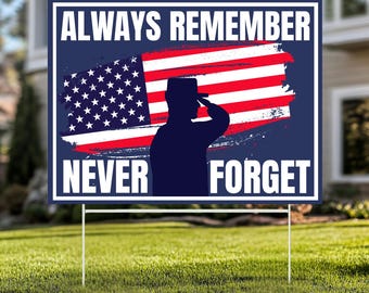 May include: A patriotic yard sign with the text "ALWAYS REMEMBER NEVER FORGET". The sign features a silhouette of a saluting soldier in front of a stylized American flag. The sign is mounted on two metal stakes and is displayed on a green lawn.