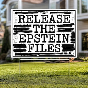 May include: A white yard sign with the text "RELEASE THE EPSTEIN FILES" in bold, black letters. The sign is on a metal stake, placed in a grassy yard. The background includes a house and trees.
