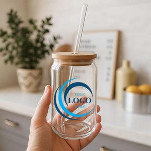 May include: A clear glass drinking cup with a bamboo lid and a glass straw. The cup has a blue and white logo that says "YOUR LOGO HERE". The cup is held in a hand.