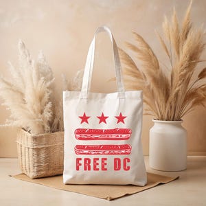May include: A white tote bag with red graphic elements. The design includes three red stars above two red sandwich illustrations, and the words "FREE DC" in red block letters. The bag has two white handles.