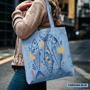 May include: Carolina blue tote bag featuring three stylized fish illustrations with yellow lemon slices and botanical accents. The word "Sardines" is printed vertically on the left side of the bag. The bag is carried by a person.